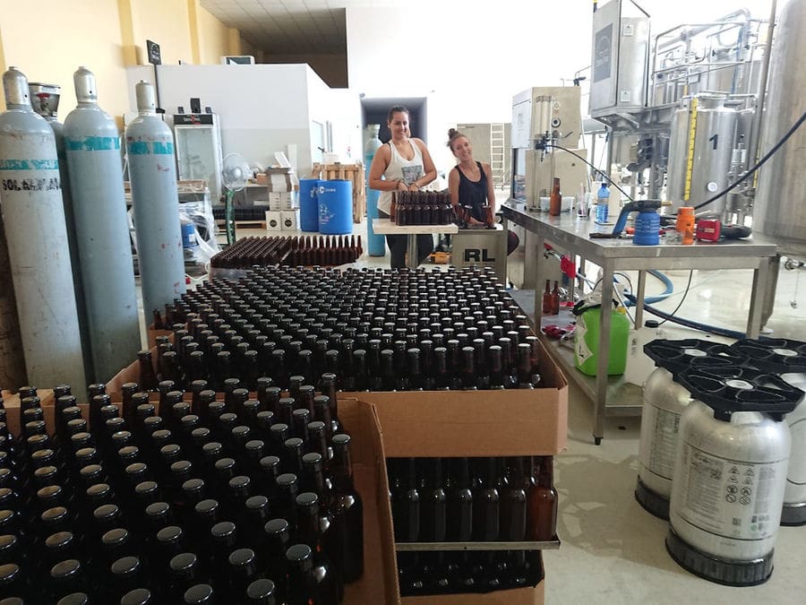 two girls smiling happily at the camera with stacked bottles in the front at 'Sknipa' plant